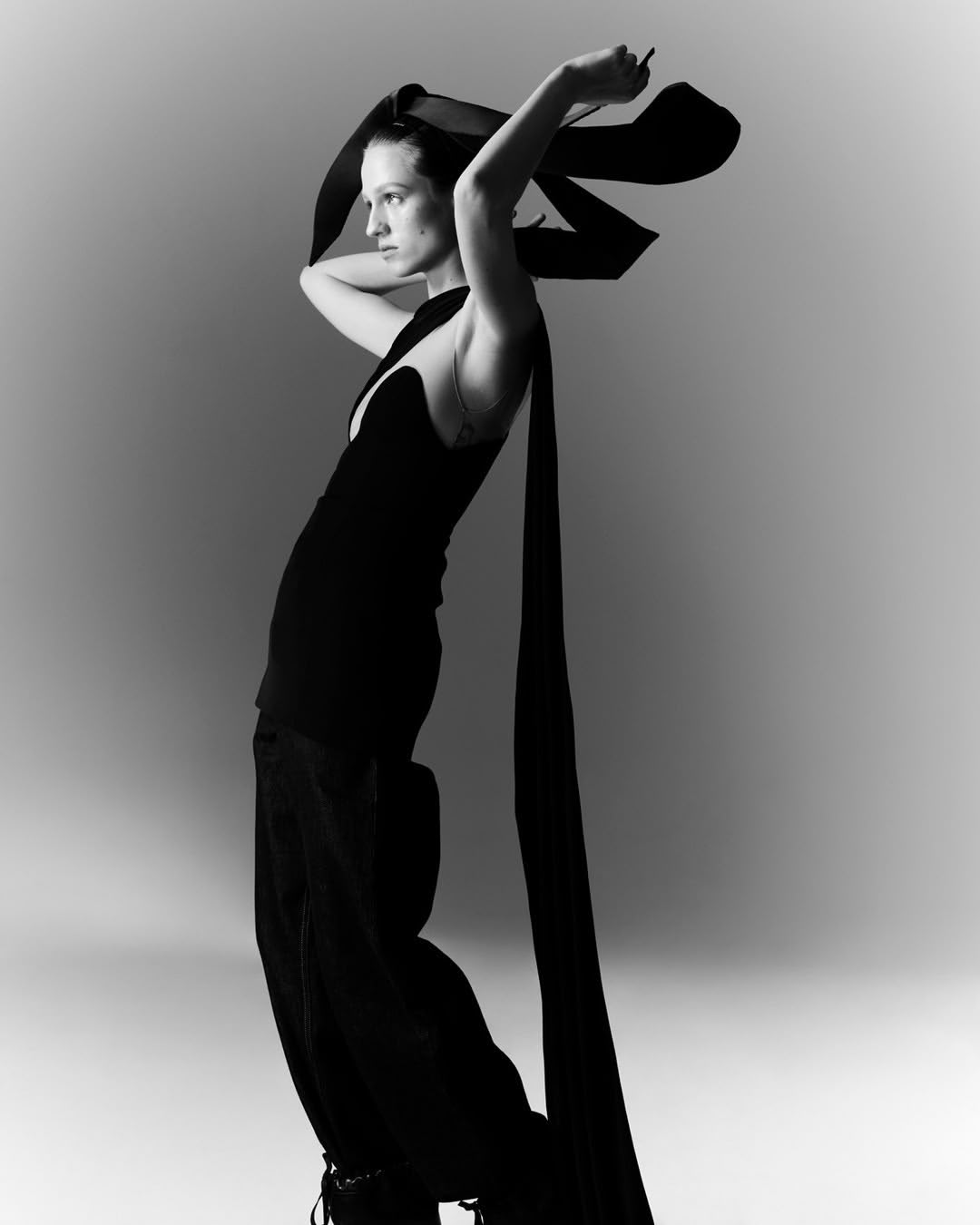 Monochrome portrait of woman in black gown and large black ribbon headpiece striking a pose in a photo studio against gray background.