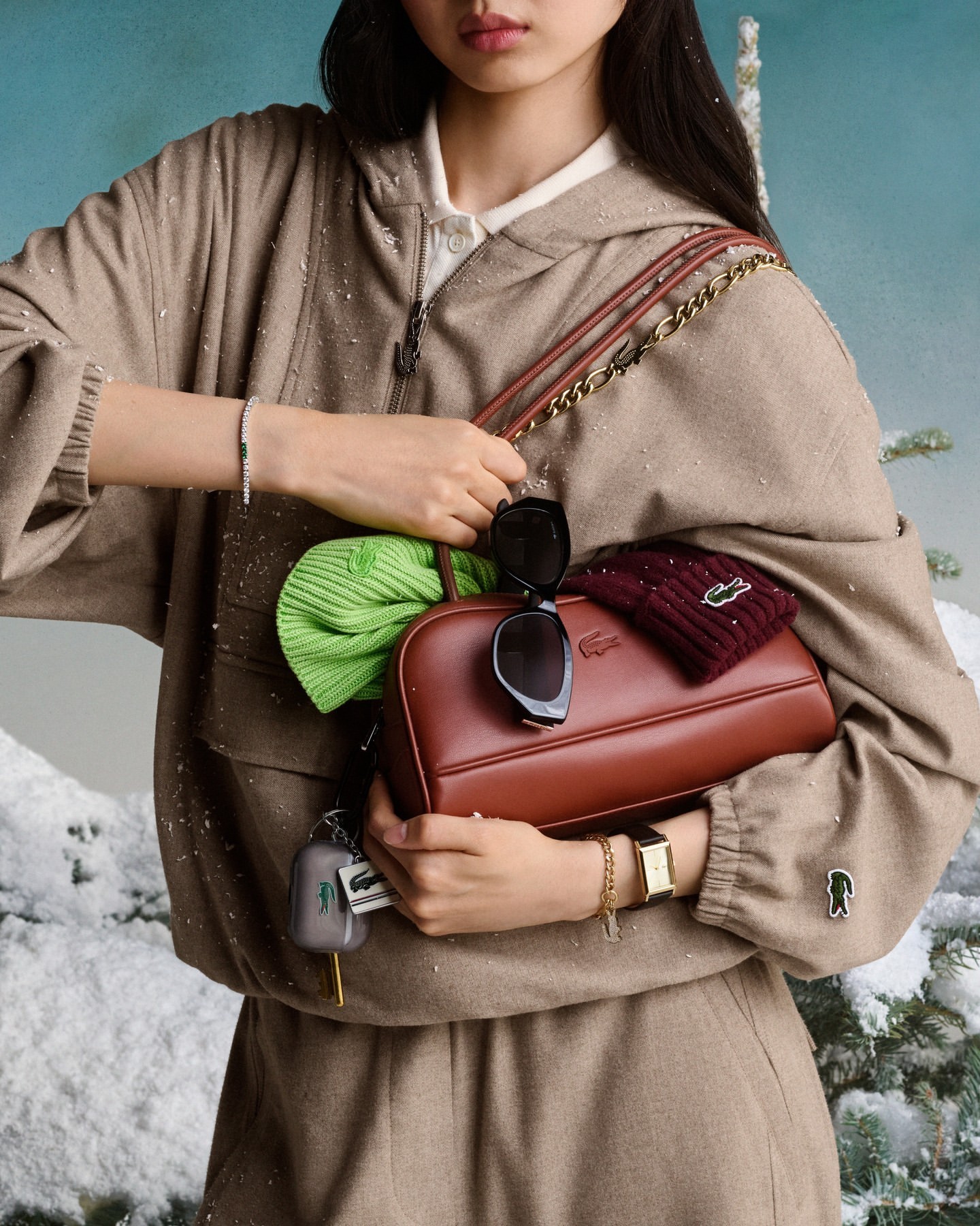 Young woman in casual clothing holds a brown leather Lacoste handbag with accessories, set against a snowy, wintry backdrop.