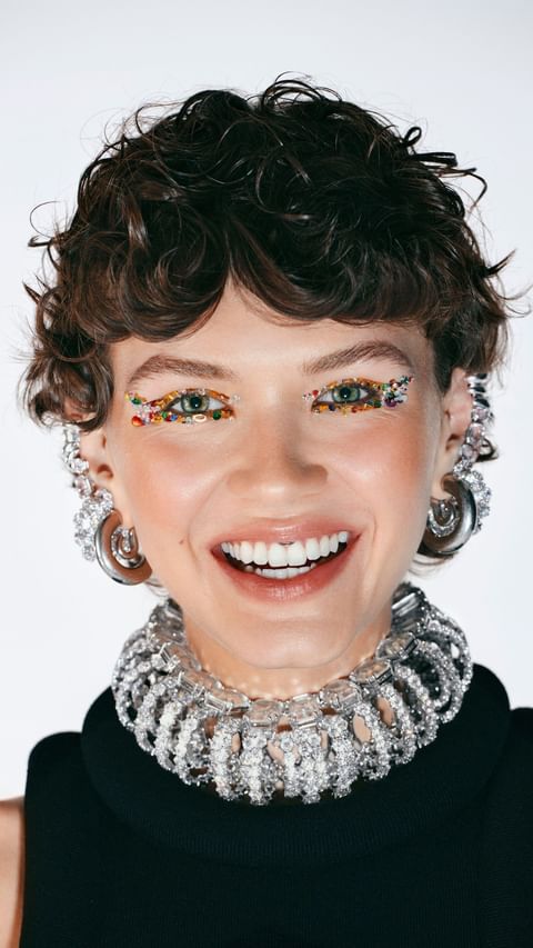 Close-up portrait of a woman with short curly brown hair, gemstone eye makeup, heavy crystal choker necklace, and silver hoop earrings.