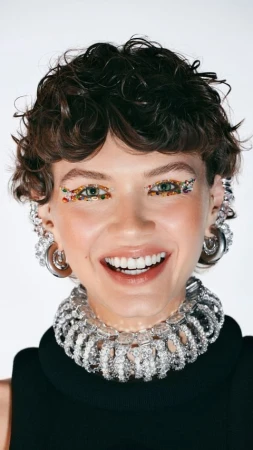 Close-up portrait of a woman with short curly brown hair, gemstone eye makeup, heavy crystal choker necklace, and silver hoop earrings.
