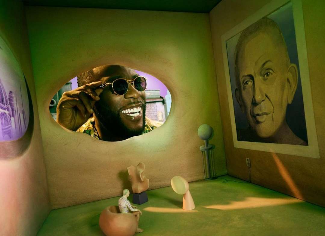 Smiling Black man in sunglasses peers through a hole in a wall of an art gallery with paintings and sculptures in green and beige hues.
