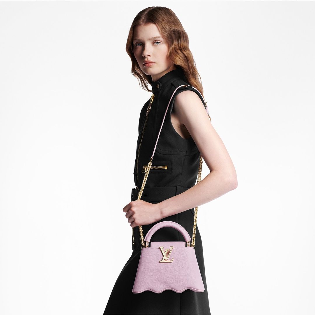 A woman in a black dress poses in a studio setting, displaying a pink leather Louis Vuitton handbag with a gold chain strap.