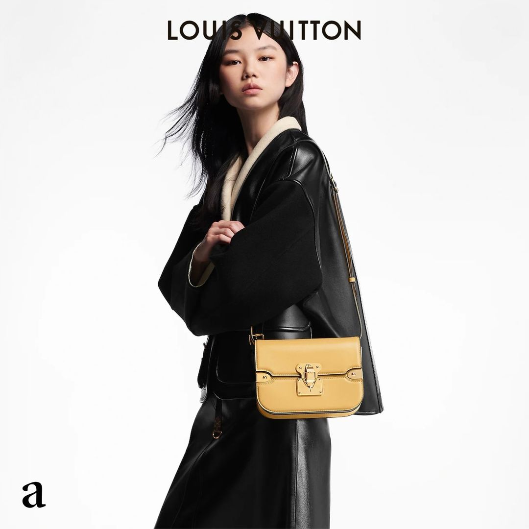 A stylish Asian woman in a black leather coat poses with a yellow leather handbag in a fashion studio shot against a white backdrop.