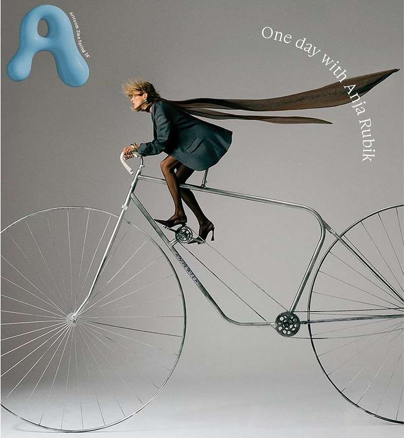 Fashion editorial: Anja Rubik in a suit jacket rides a vintage bicycle with oversized wheels. Gray backdrop with magazine graphics.