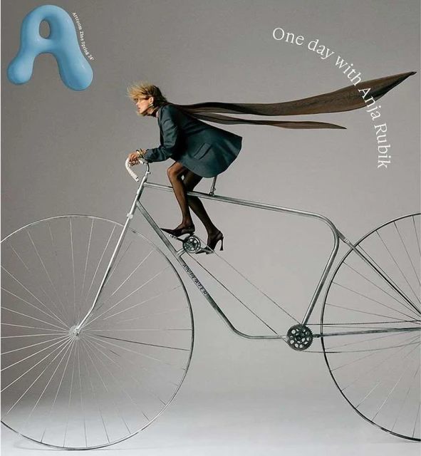 Fashion editorial: Anja Rubik in a suit jacket rides a vintage bicycle with oversized wheels. Gray backdrop with magazine graphics.