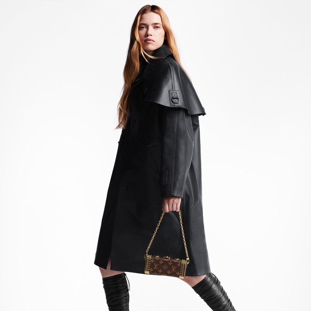 Blonde woman in a black trench coat holding a Louis Vuitton box bag in a studio setting, fashionable and chic, with high-fashion appeal.