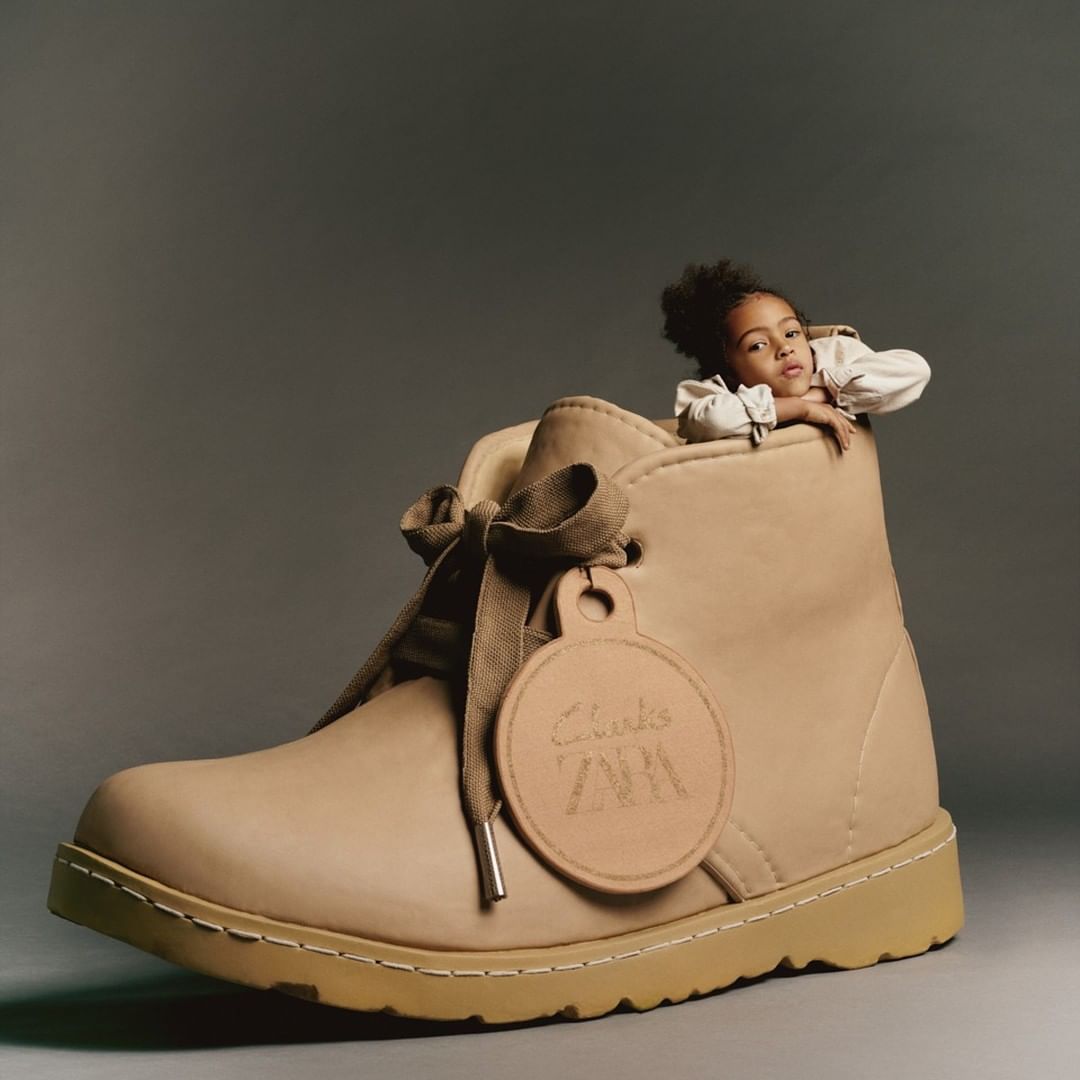 A young child is peeking out of the top of an oversized Clarks brand shoe, with the brand logo visible on an attached tag.