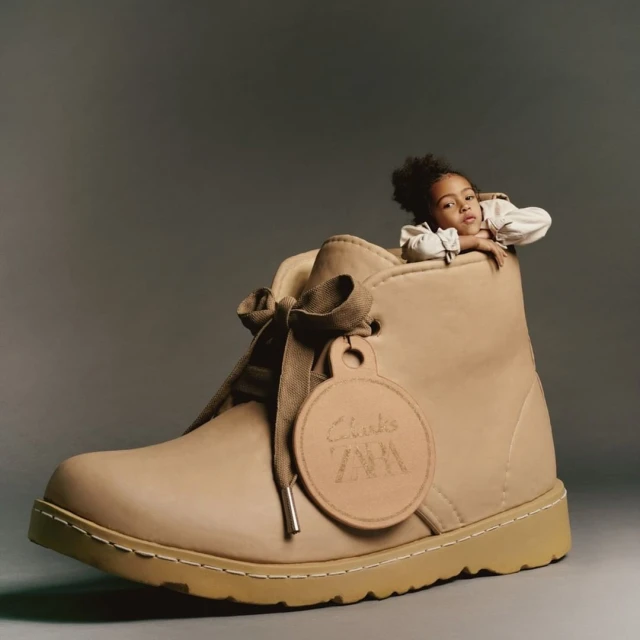A young child is peeking out of the top of an oversized Clarks brand shoe, with the brand logo visible on an attached tag.