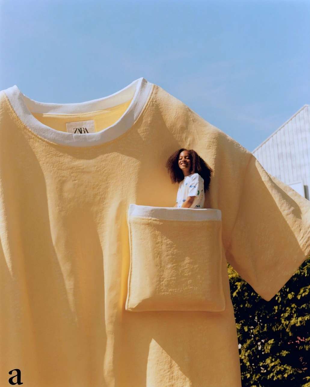A young adult Black woman is pictured inside the pocket of an oversized yellow t-shirt against a backdrop of blue sky and green foliage.