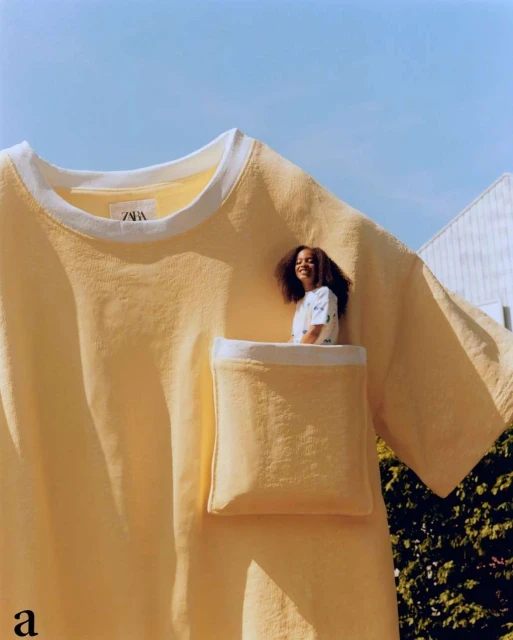 A young adult Black woman is pictured inside the pocket of an oversized yellow t-shirt against a backdrop of blue sky and green foliage.