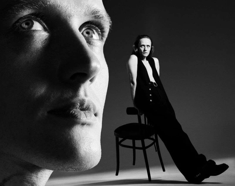 Black and white portrait featuring a close-up of a fair-skinned person's face alongside a person in a jumpsuit posing on a chair.