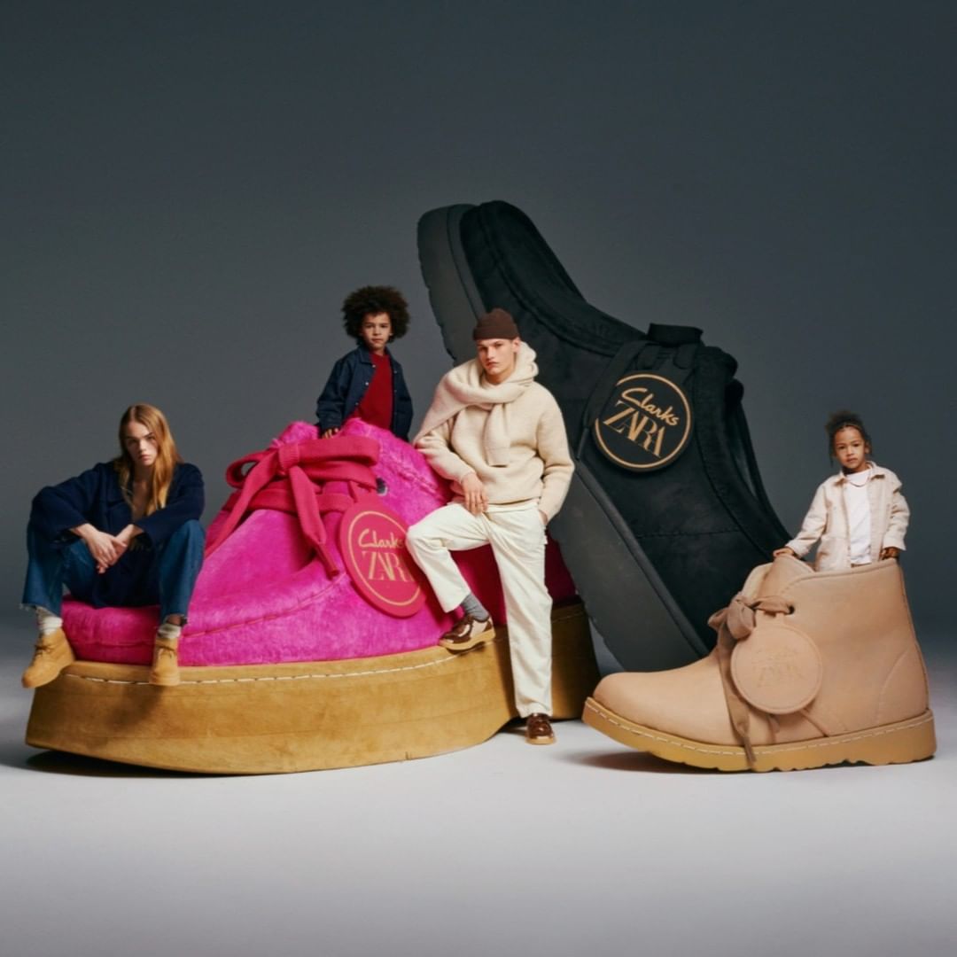 People sit on and stand by giant shoes for Clarks and Zara clothing and shoe collaboration against a gray studio background.