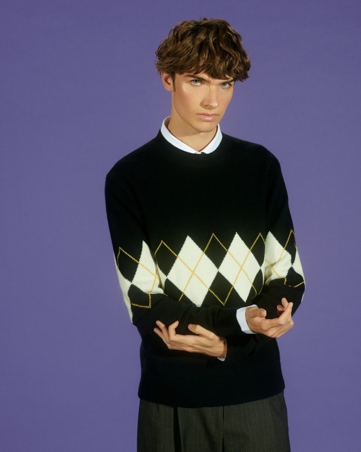 Studio shot of a young adult man in a black and white argyle sweater and collared shirt, posing against a solid purple backdrop.