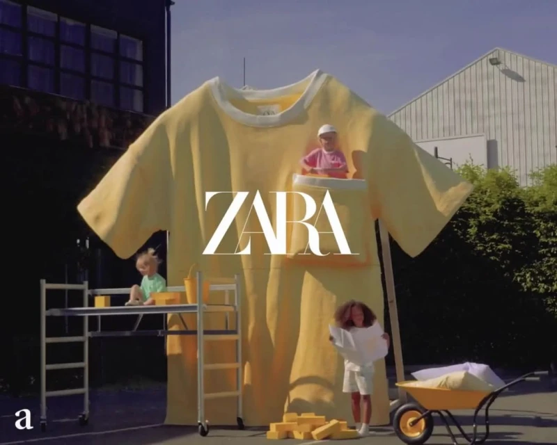 Child-sized T-shirt product display with two kids on scaffolding and holding blueprints, plus another doll figure in pocket, outdoors.