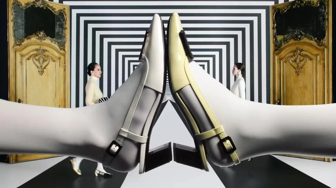 Eye-level studio shot of two women standing near decorative doors and a geometric striped wall, with a focus on a pair of shoes.