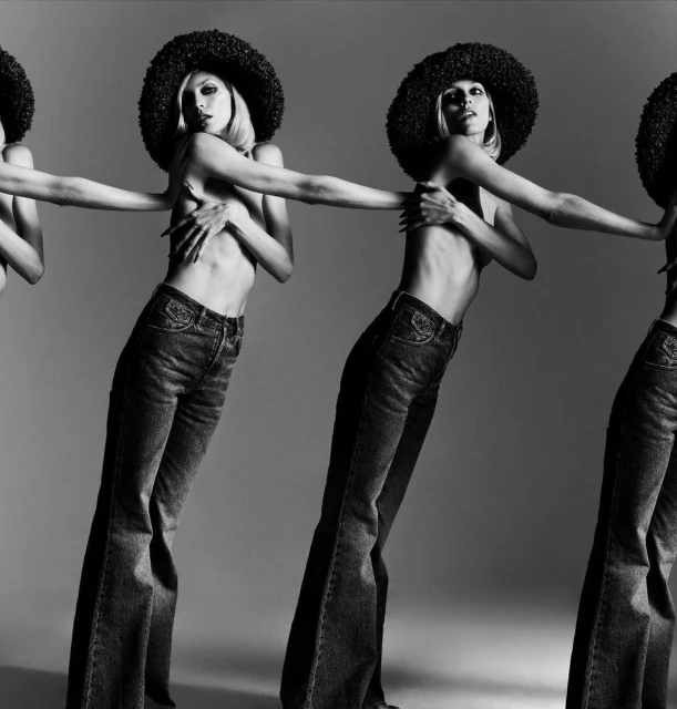 Black and white studio shot of a row of three topless female models in jeans and afro wigs, arms linked, fashion editorial.