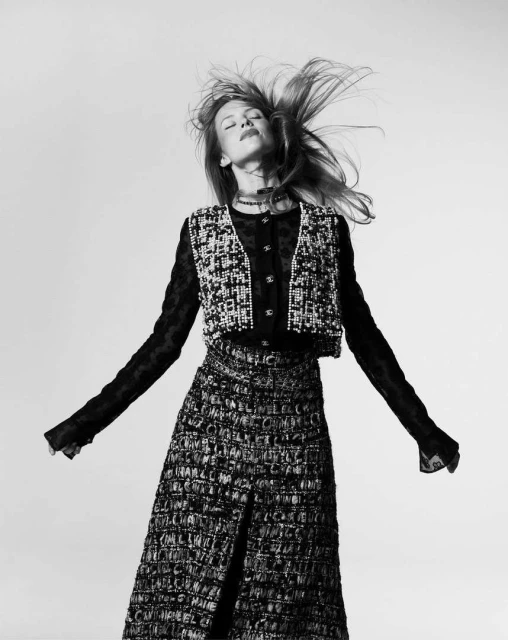 A woman in a tweed skirt and vest poses with her arms outstretched and her hair blowing in a black and white studio shot.
