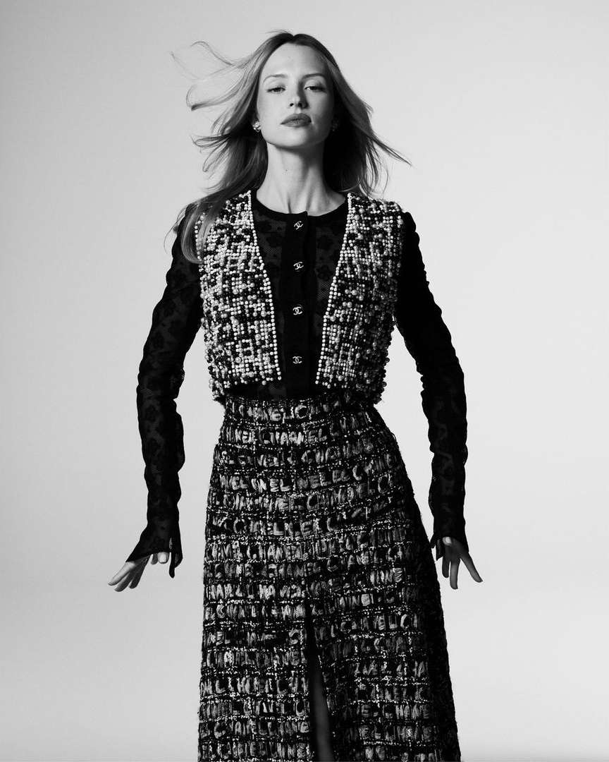Monochrome fashion shot of a model in an embellished tweed skirt, vest, and long-sleeved top, posing against a bright backdrop.