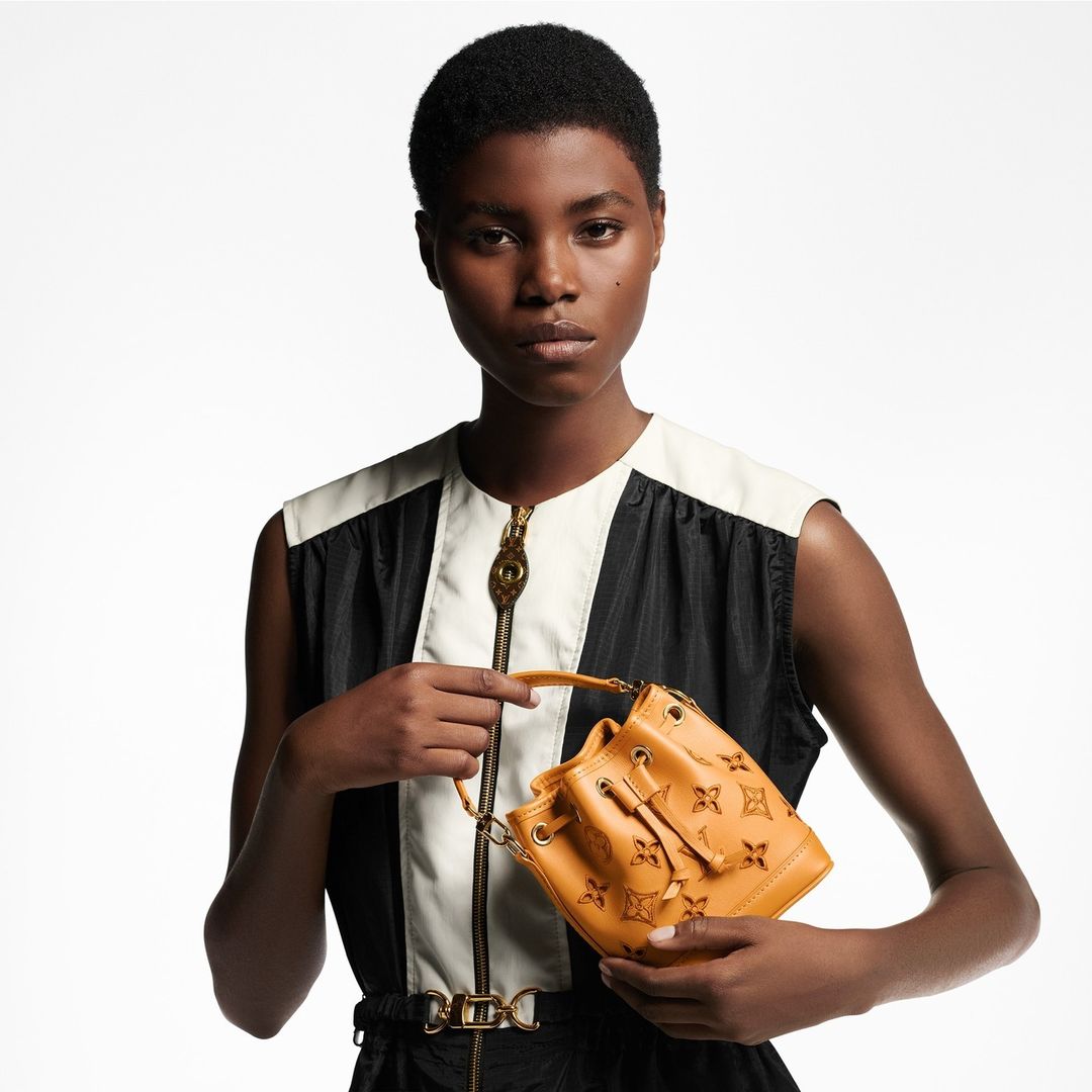 Adult female model presents a tan leather Louis Vuitton bucket bag against a bright white background in a studio setting.