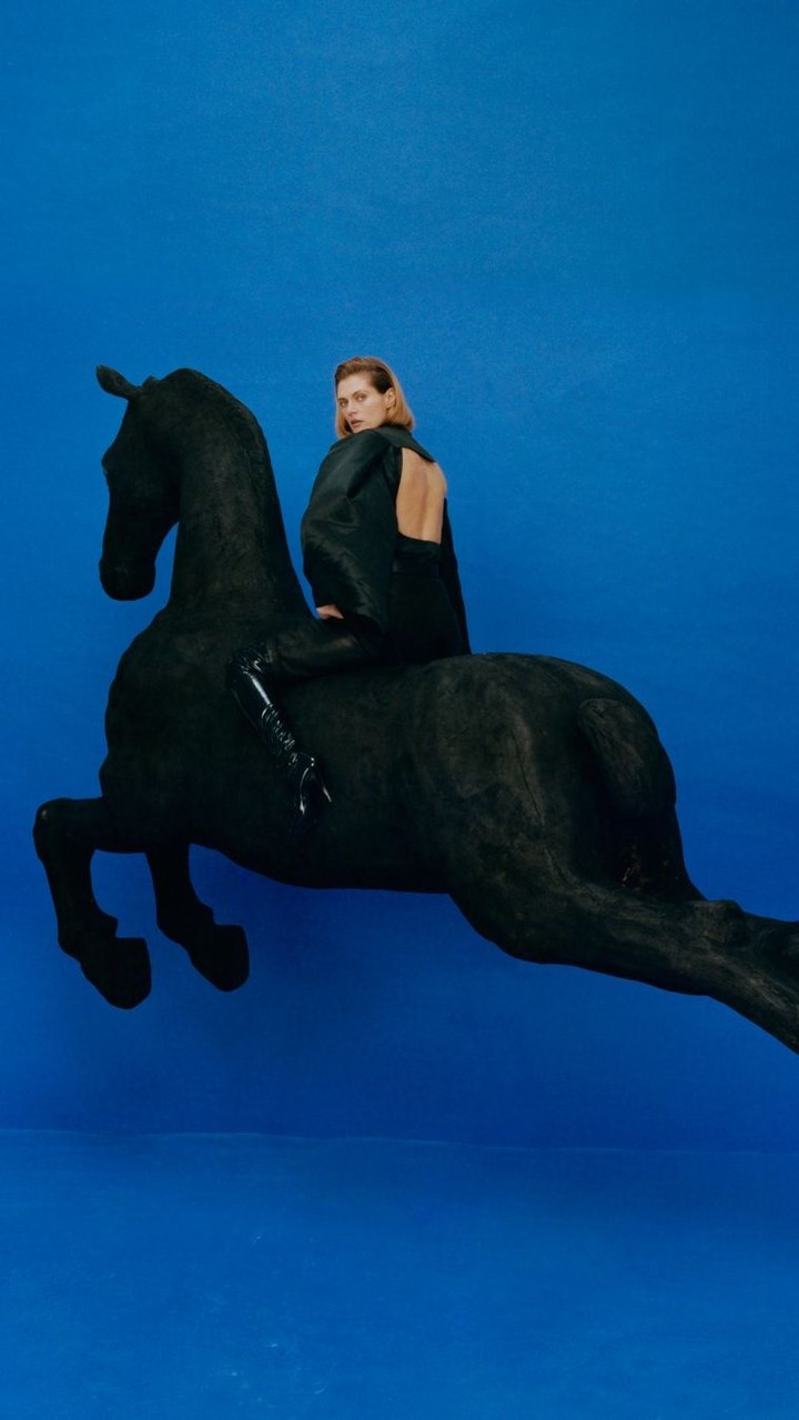 A blond woman in a black dress sits atop a black horse sculpture against a bright blue backdrop, creating a striking studio fashion image.