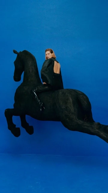 A blond woman in a black dress sits atop a black horse sculpture against a bright blue backdrop, creating a striking studio fashion image.