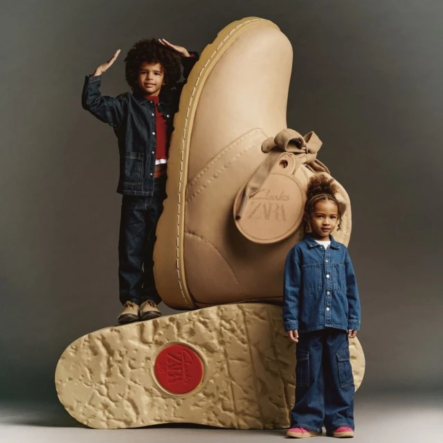Two children, a boy and a girl, pose next to a giant Clarks shoe for a Clarks and Zara brand collaboration advertisement in a studio.