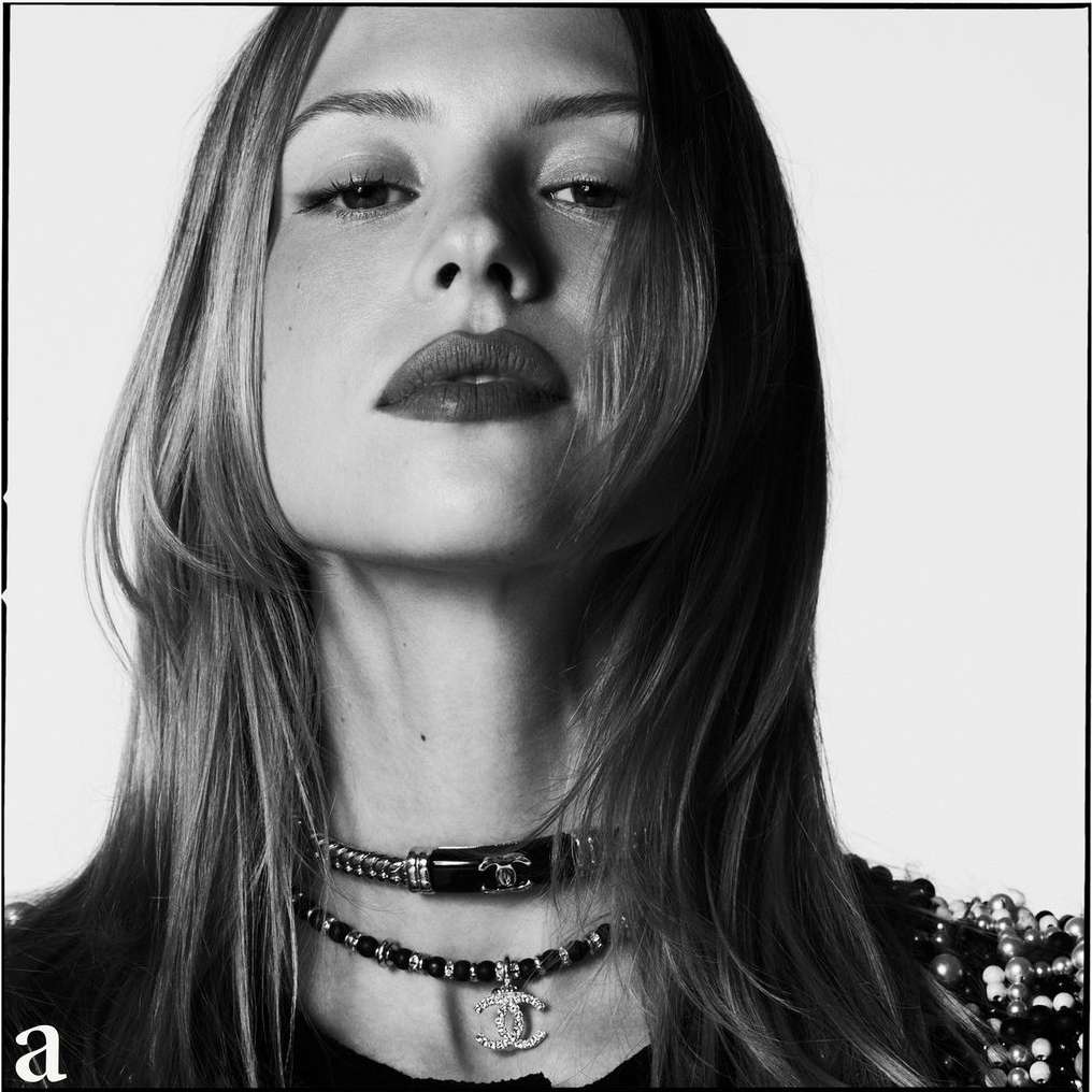 Black and white close-up studio shot of a blonde adult female model wearing two Chanel choker necklaces, looking directly at the camera.