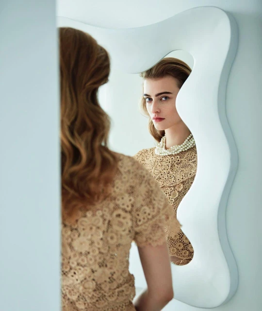 Adult woman wearing lace top and pearl necklace gazes at her reflection in a modern white mirror against a white wall.