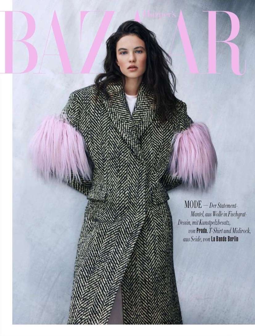 Fashion editorial shot of a woman wearing a black and white herringbone coat with pink fur sleeves against a light pink backdrop.