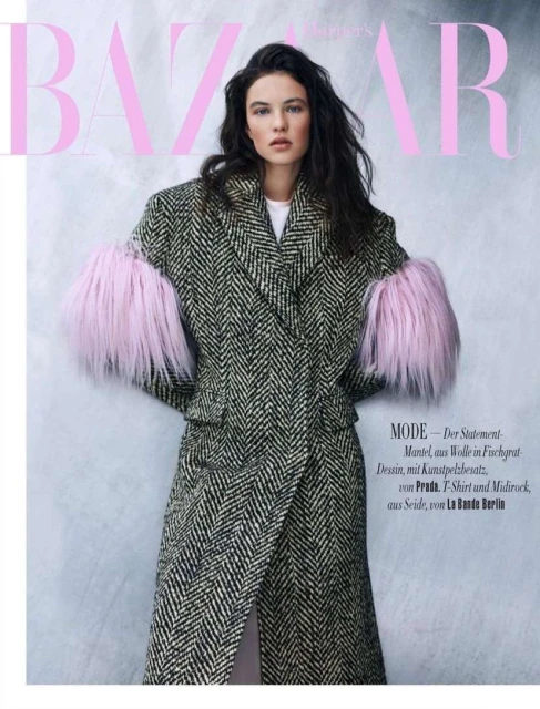 Fashion editorial shot of a woman wearing a black and white herringbone coat with pink fur sleeves against a light pink backdrop.