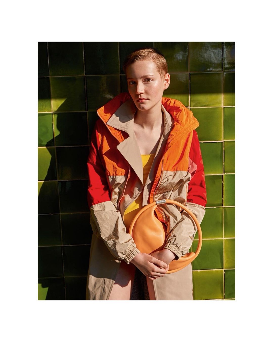 Adult female poses indoors near a green tiled wall, wearing a tan and red jacket and holding an orange round purse. Fashion shot.