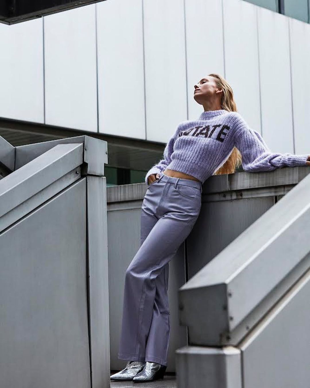 Adult woman in sweater and flared pants posing on stairs in an outdoor setting with a neutral palette and geometric architecture.