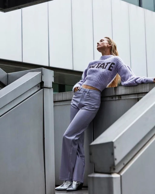 Adult woman in sweater and flared pants posing on stairs in an outdoor setting with a neutral palette and geometric architecture.