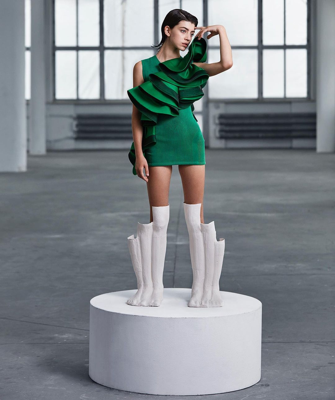 A fashion shot of a young woman posing on a white platform wearing a short green dress with large ruffles and sculptural white boots.