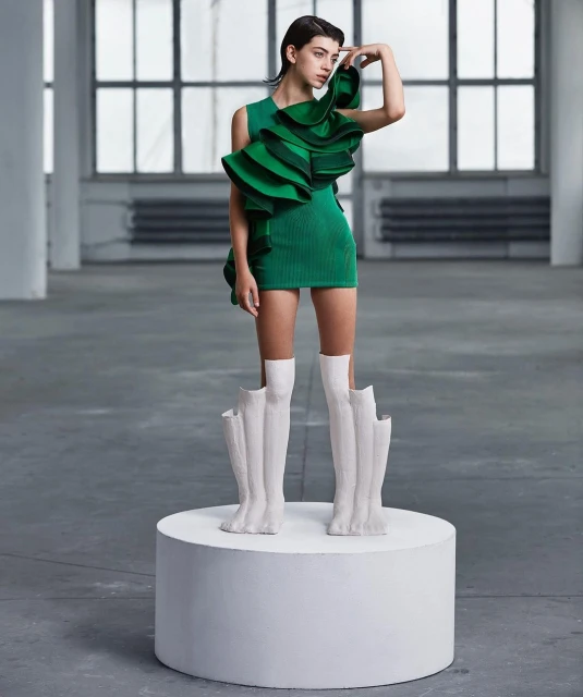 A fashion shot of a young woman posing on a white platform wearing a short green dress with large ruffles and sculptural white boots.