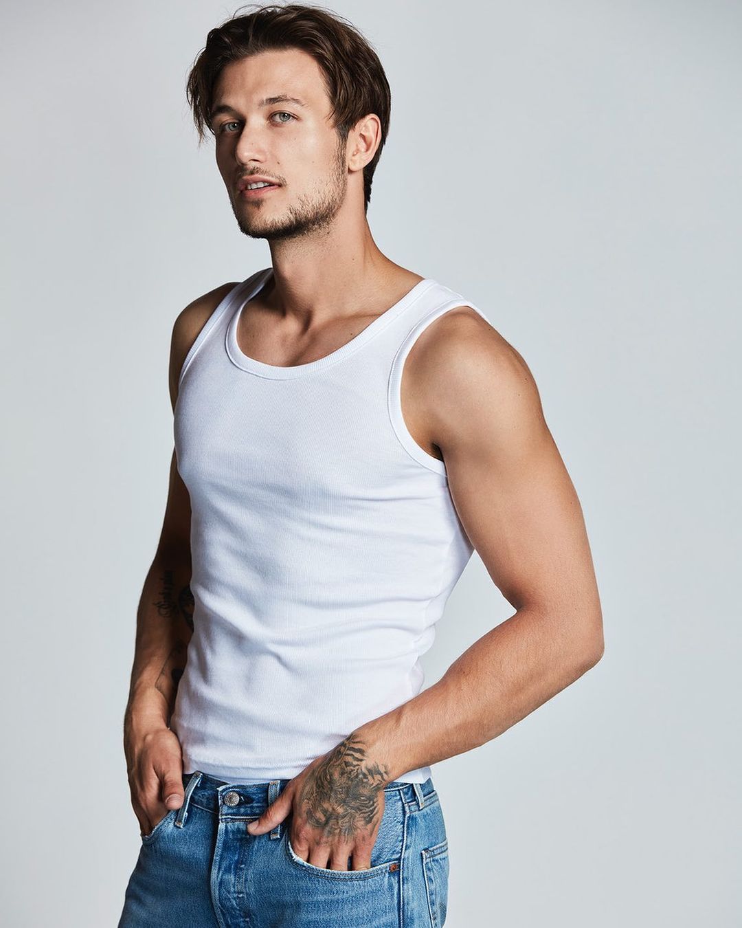 Man wearing a white tank top and blue jeans posing against a clean white background