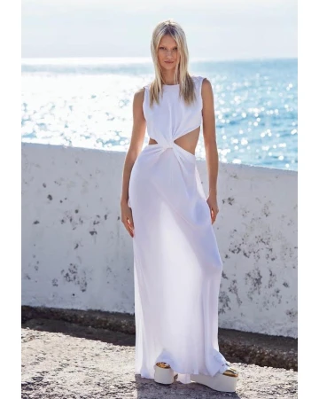 Blonde woman in a white cut-out maxi dress standing on a coastal wall overlooking the sea by Marcin Tyszka