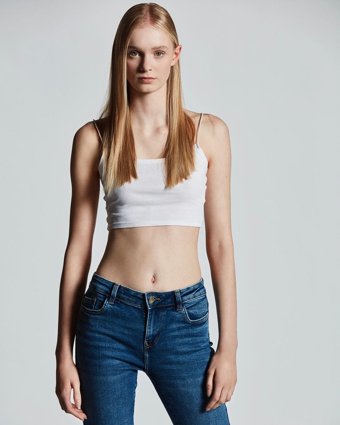 Portrait of a blonde female model in a white crop top and blue jeans against a grey seamless background.