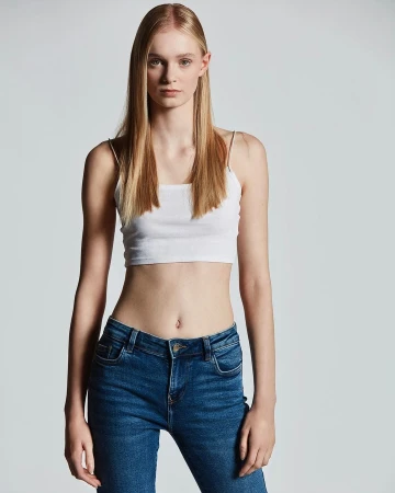 Portrait of a blonde female model in a white crop top and blue jeans against a grey seamless background.