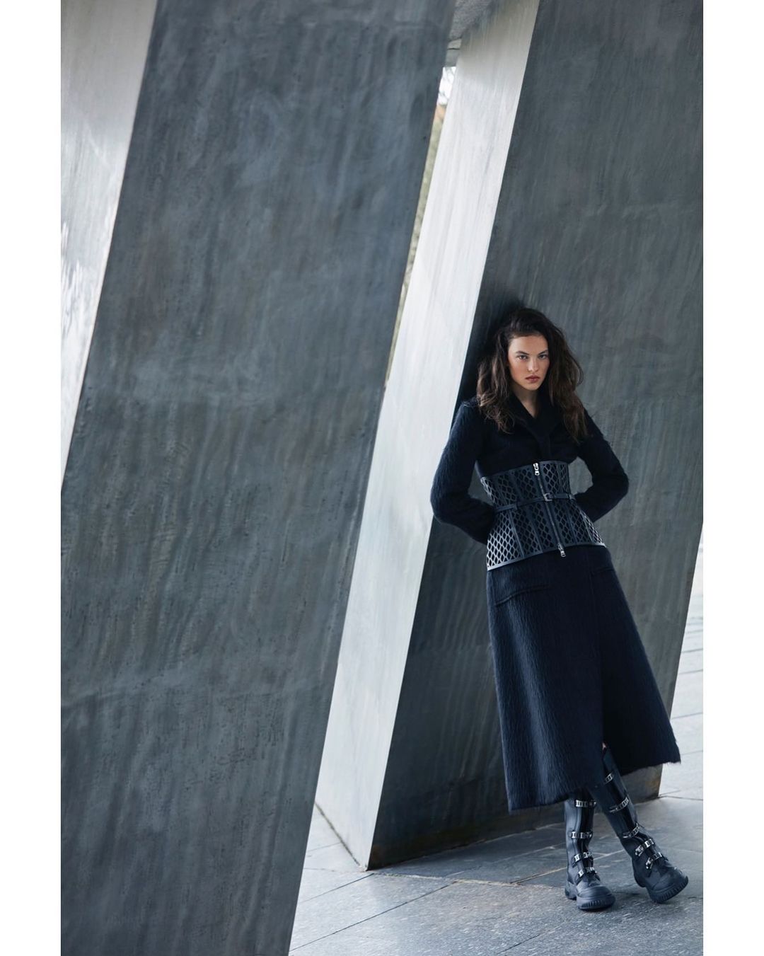 Woman in a black coat and corset posing against a concrete wall, showcasing fashion and editorial style with modern architecture.
