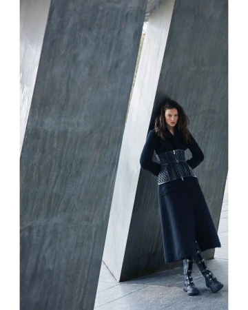 Woman in dark coat and patterned corset leaning against large concrete pillars in Milan by Marcin Tyszka