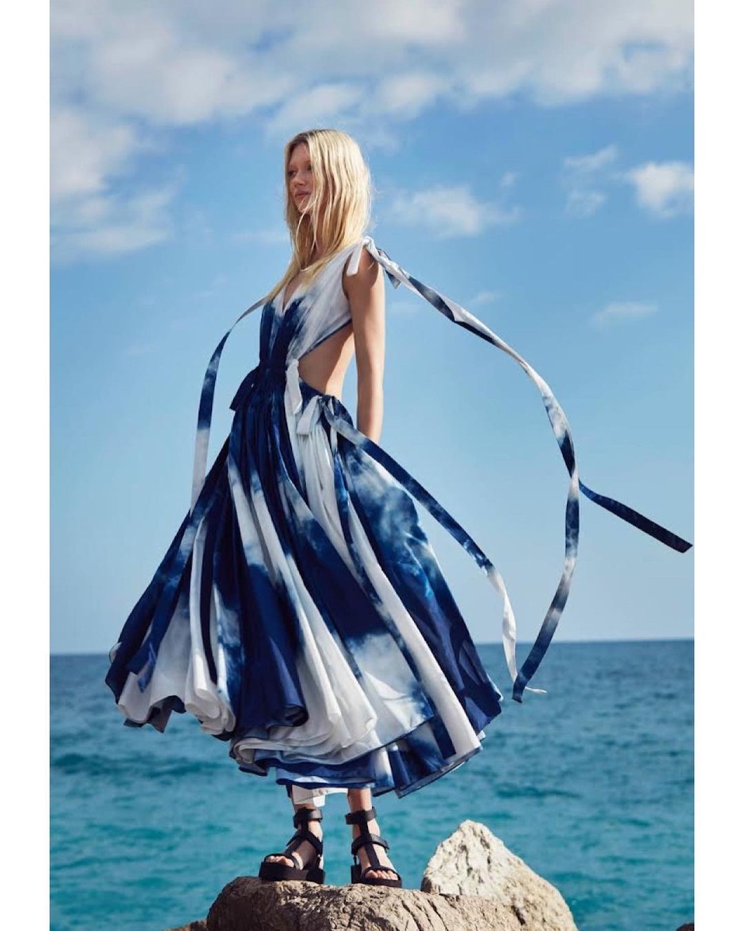 A woman in a blue and white tie-dye dress stands on rocks by the sea with a cloudy blue sky backdrop, showcasing fashion and style.