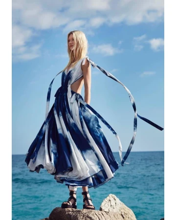 A woman in a blue and white tie-dye dress stands on rocks by the sea with a cloudy blue sky backdrop, showcasing fashion and style.
