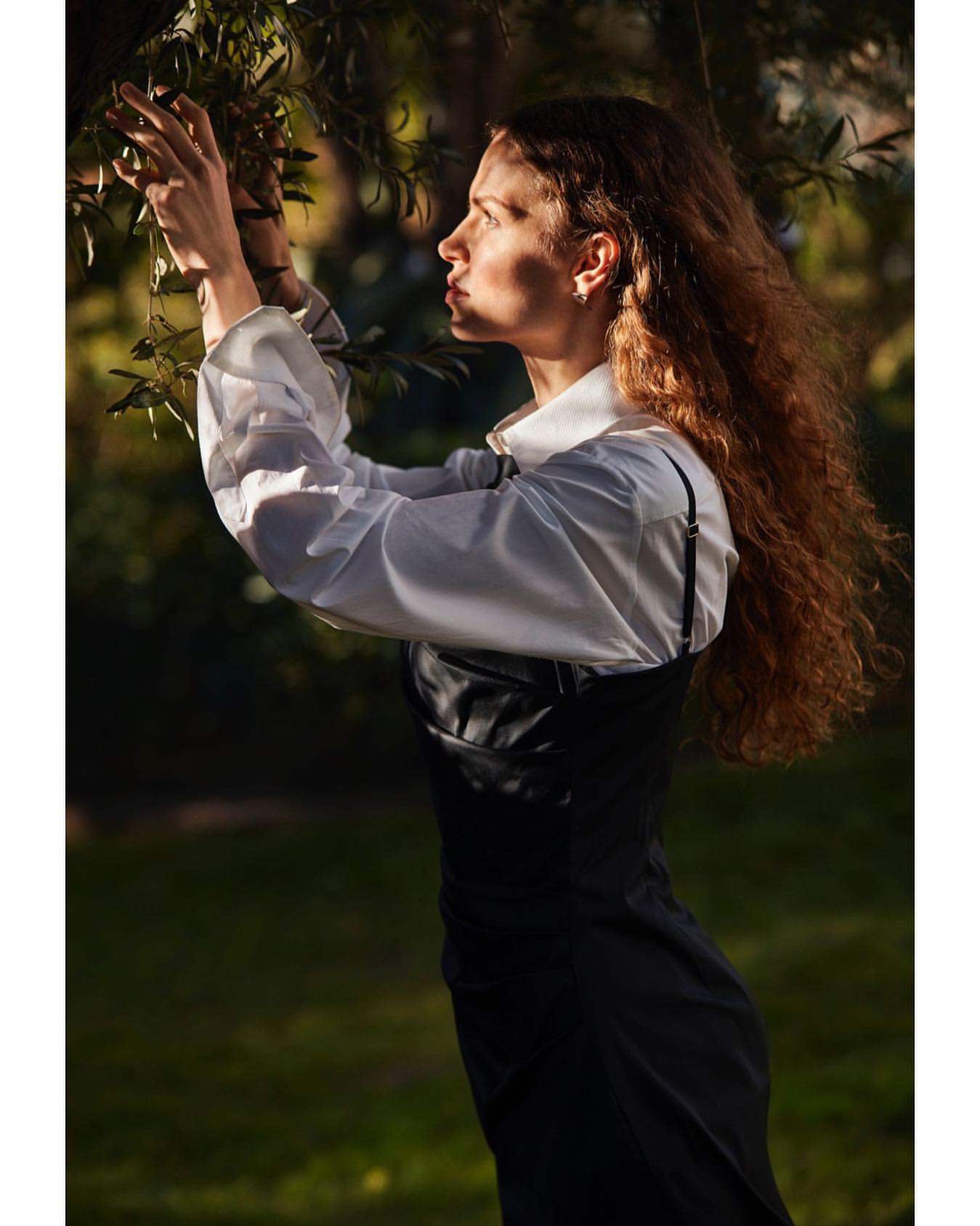 Profile of a woman with long curly brown hair wearing a white shirt and black dress, reaching for tree branches outdoors.