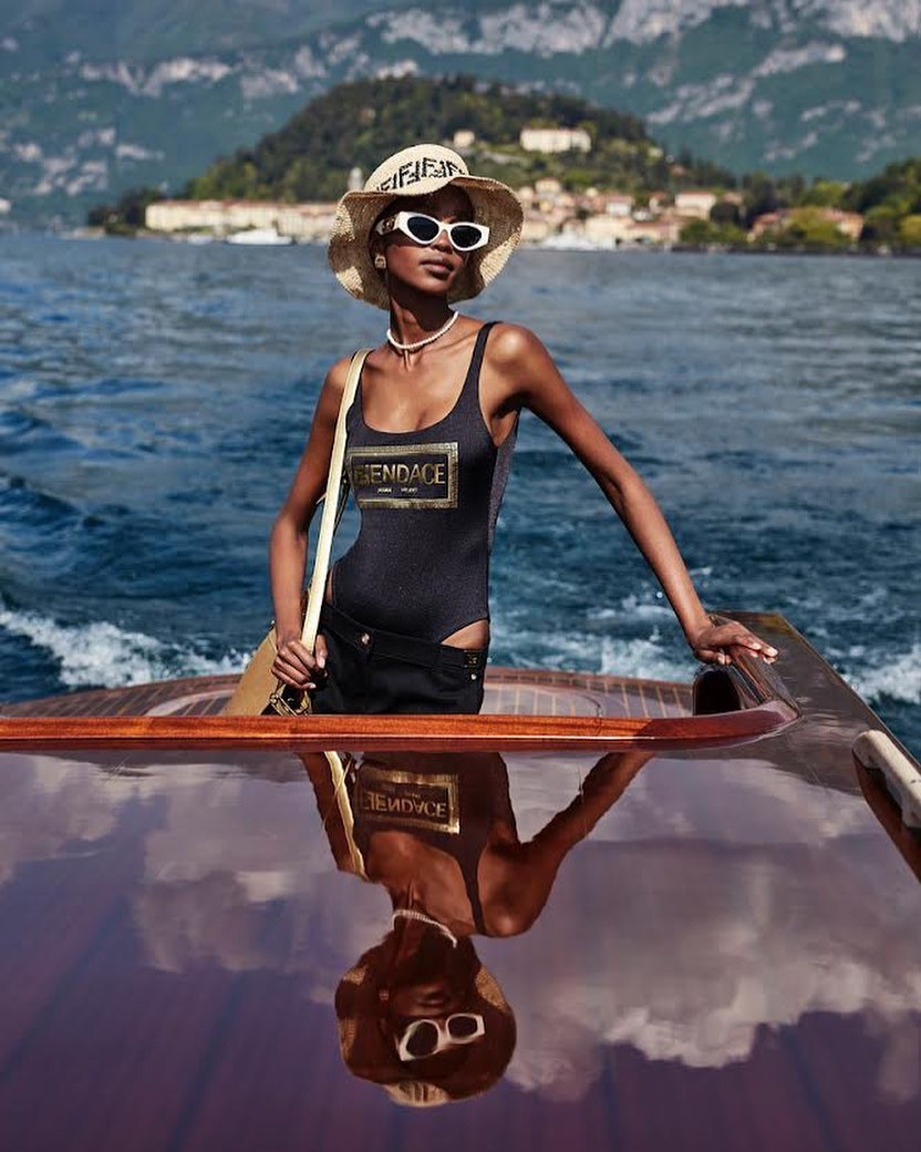 Dark-skinned woman in a bathing suit and straw hat poses on a wood boat on Lake Como with a landscape backdrop on a sunny day.
