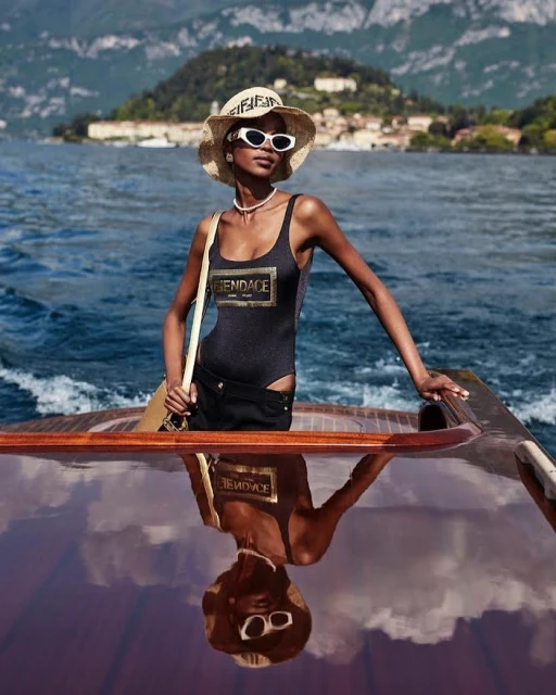 Dark-skinned woman in a bathing suit and straw hat poses on a wood boat on Lake Como with a landscape backdrop on a sunny day.