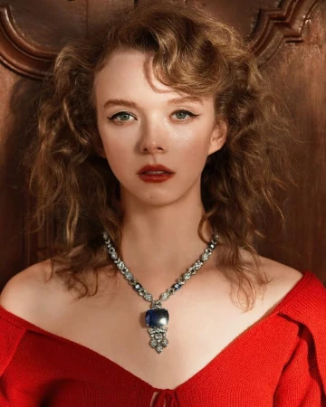 Close-up portrait of an adult woman with curly brown hair, wearing a red top and a large blue sapphire necklace, against a wood background.