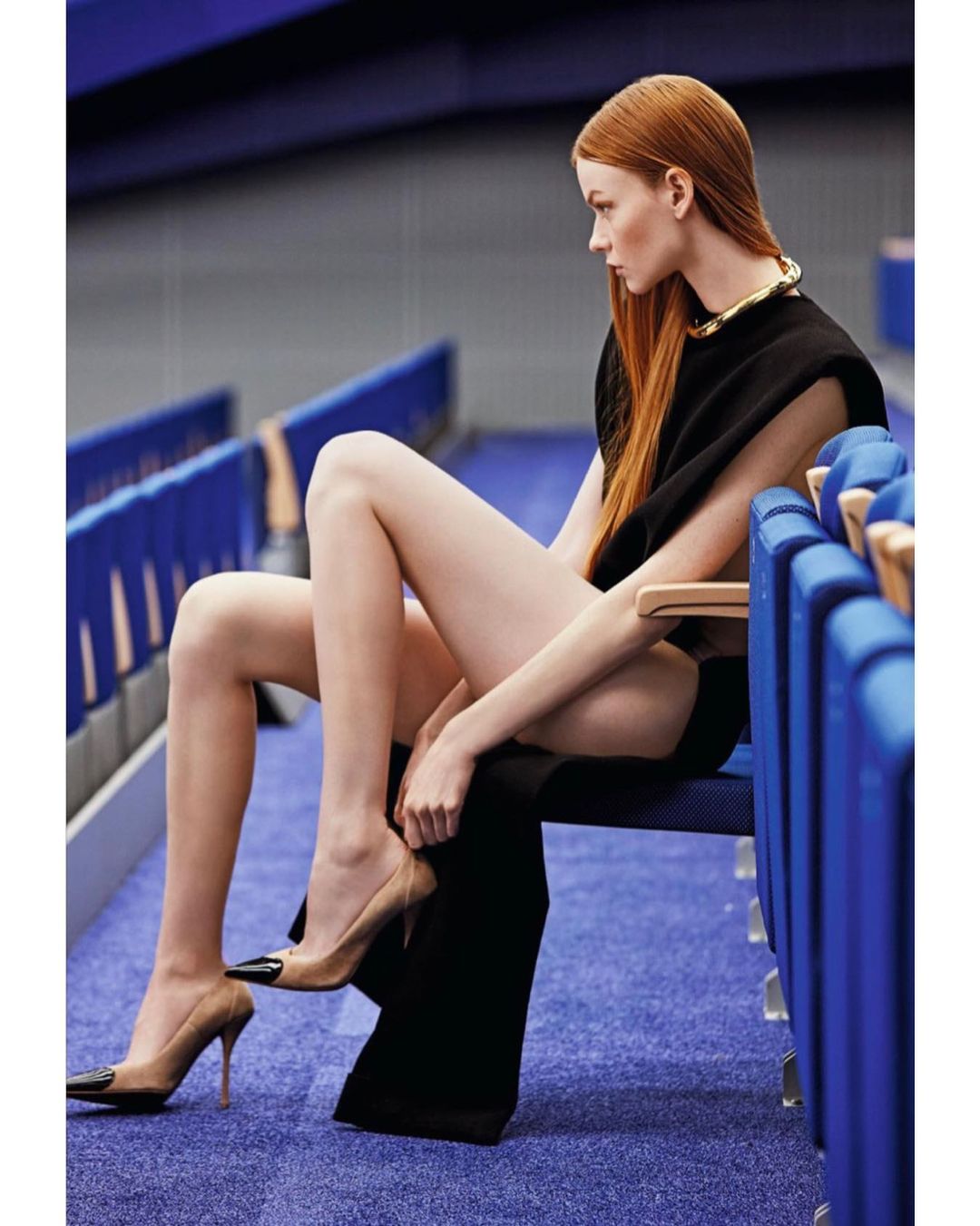 A redhead fashion model is sitting in an indoor stadium wearing a black dress, gold necklace, and high heels in a medium shot.