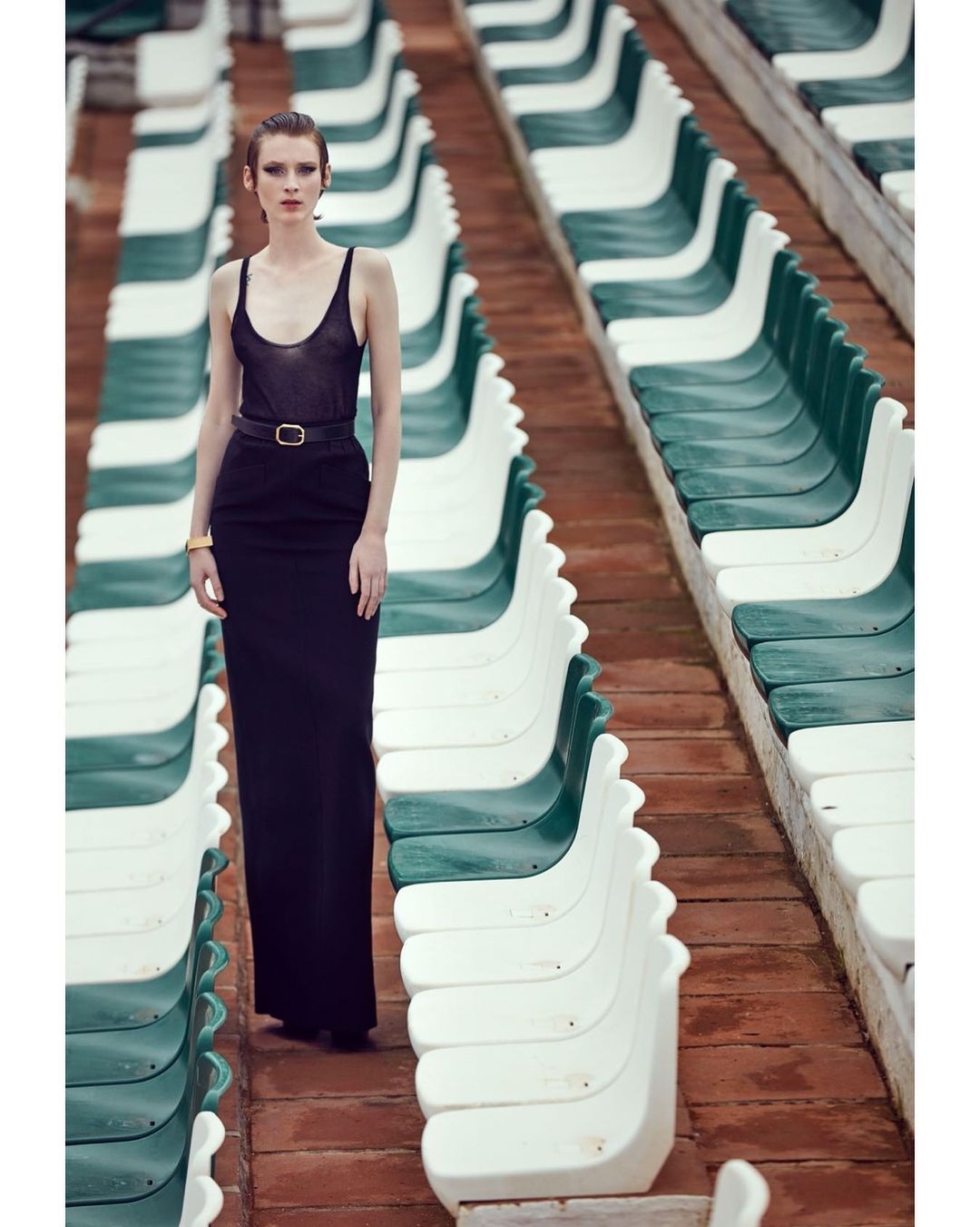 A solitary woman wearing a sleek black dress stands in an empty stadium, fashionably posing amongst rows of empty green and white seats.