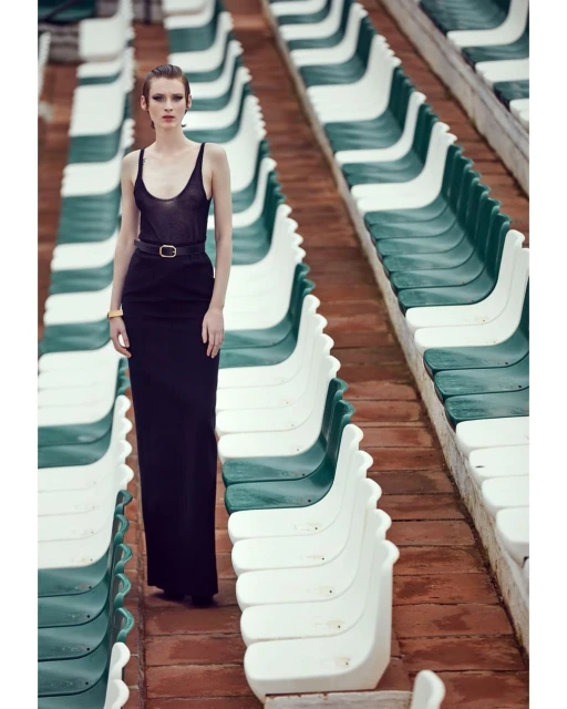 A solitary woman wearing a sleek black dress stands in an empty stadium, fashionably posing amongst rows of empty green and white seats.